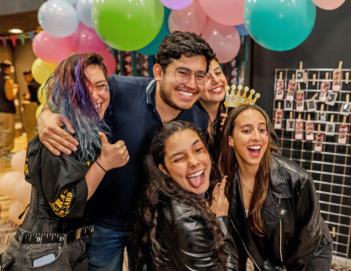 A group of five people are happily posing for a photo at a festive gathering. They are surrounded by colorful balloons and a string of photos in the background. The person in front is sticking out their tongue, and another is wearing a paper crown.